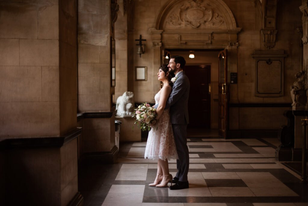 Bride and groom standing close together in a stone town hall corridor, the bride holding a pale pink bouquet