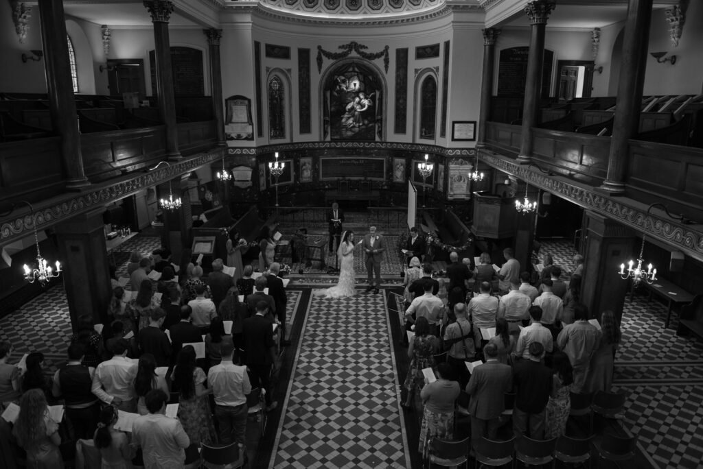 Wedding ceremony seen from the gallery of a London church, guests standing in the pews, black and white photograph