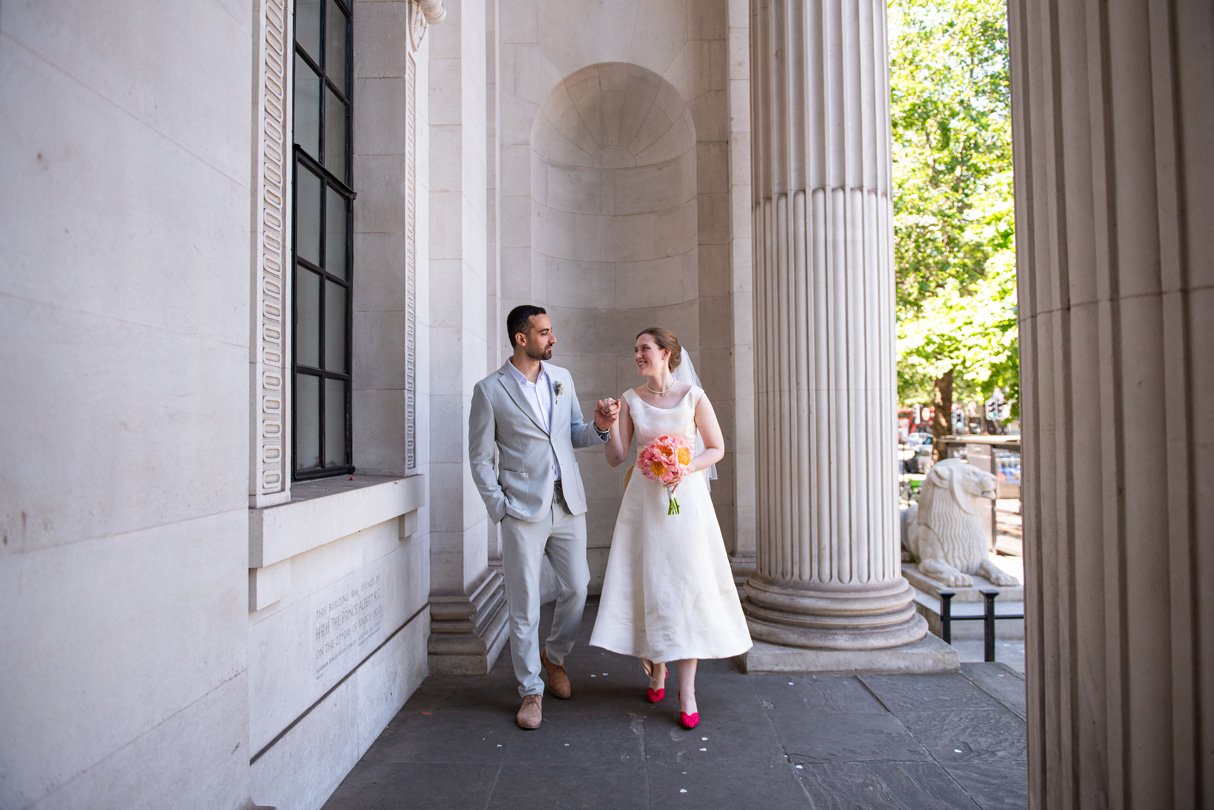 Newlyweds walking hand in hand under the columns of Old Marylebone Town Hall, the bride carrying a coral bouquet