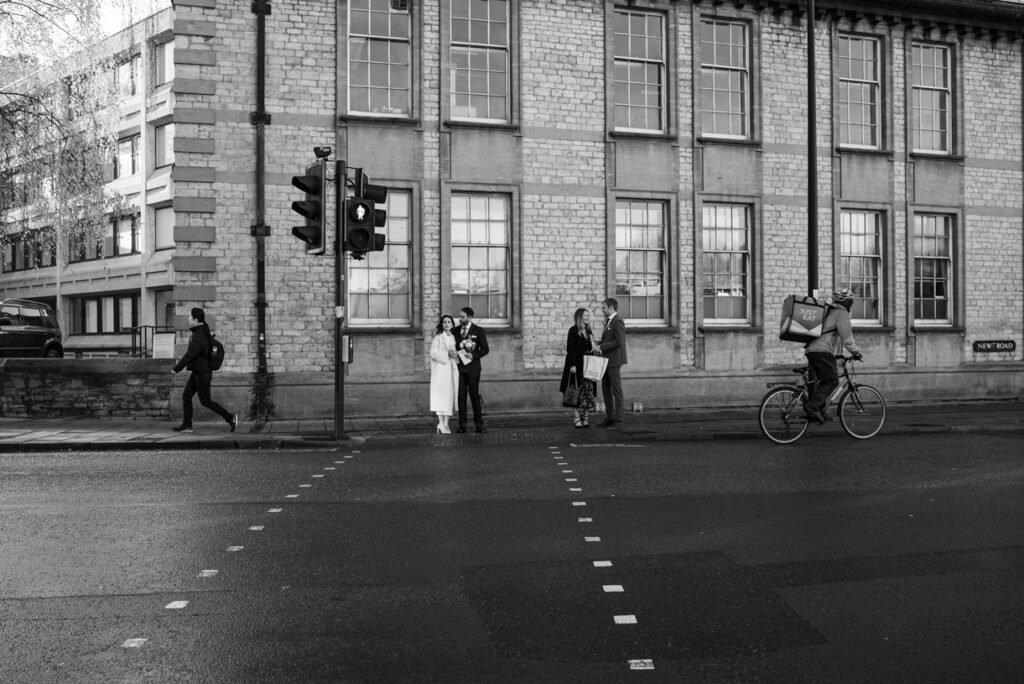 Bride and groom pausing on a city street corner as a cyclist and passers-by go about their day, black and white photograph