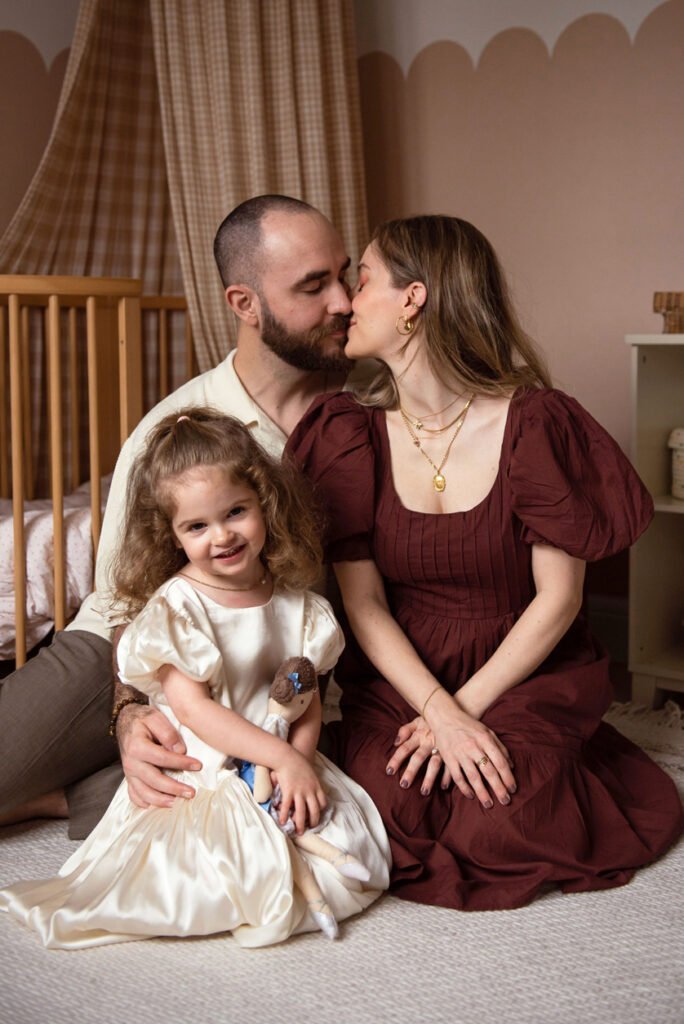 Parents kissing on their bedroom floor while their small daughter leans against them