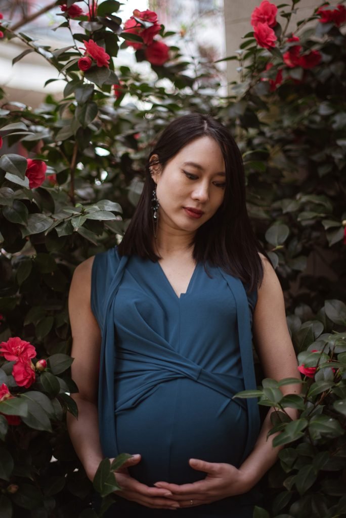 Expectant mother holding her bump in front of a camellia bush in flower