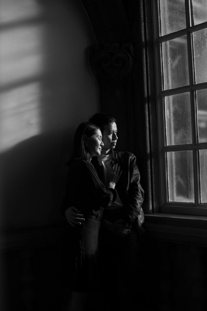 Couple in near darkness beside a tall window, lit only by the light from outside