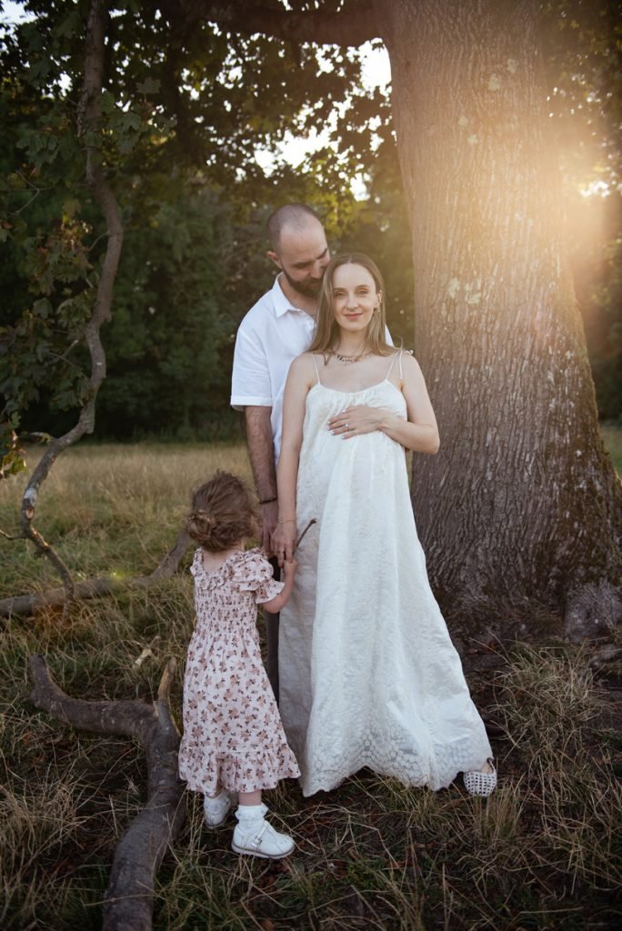 Expectant couple and their small daughter under a tree at sunset