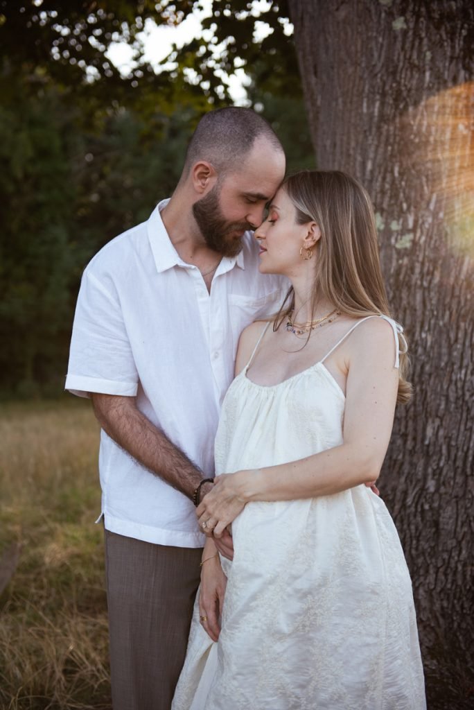 Expectant couple standing close beside a tree in a meadow