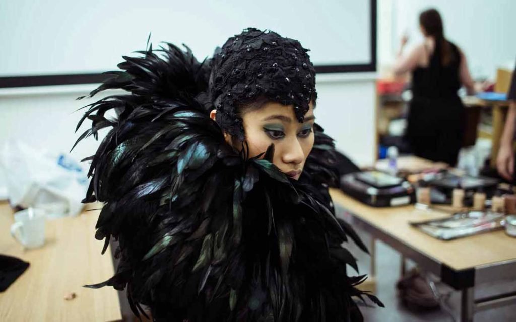 Performer in a black feathered headdress and cape backstage