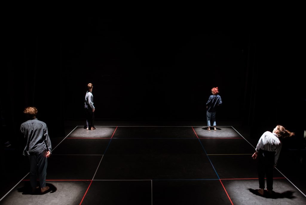 Dancers spaced across a dark stage marked out with lines of light