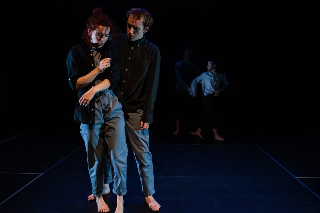 Dancers holding each other upright at the front of a dark stage