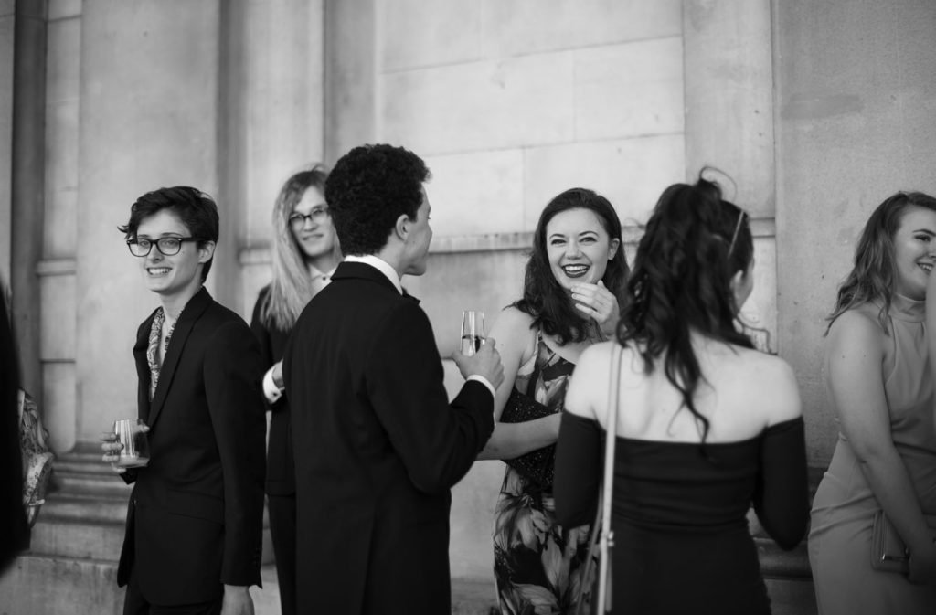 Guests in black tie talking and laughing in a stone hall, black and white photograph