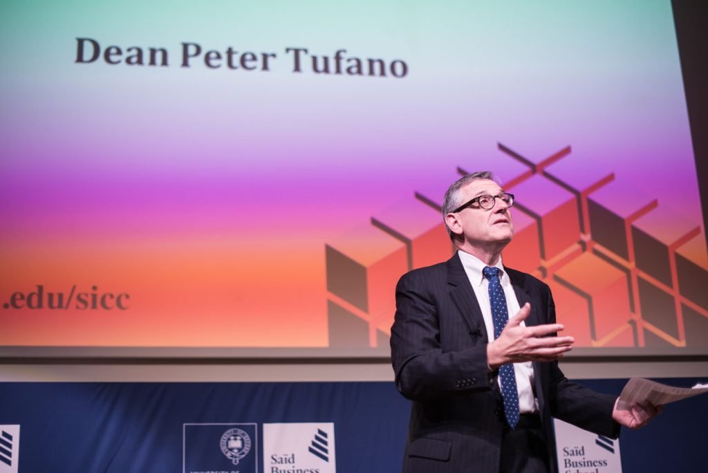 Speaker on stage at a business school conference, title slide behind him