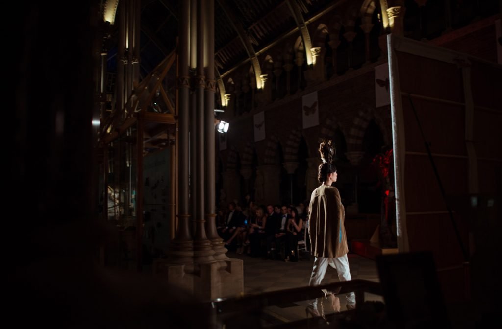 Model on a runway in a vaulted hall, audience seated along the sides