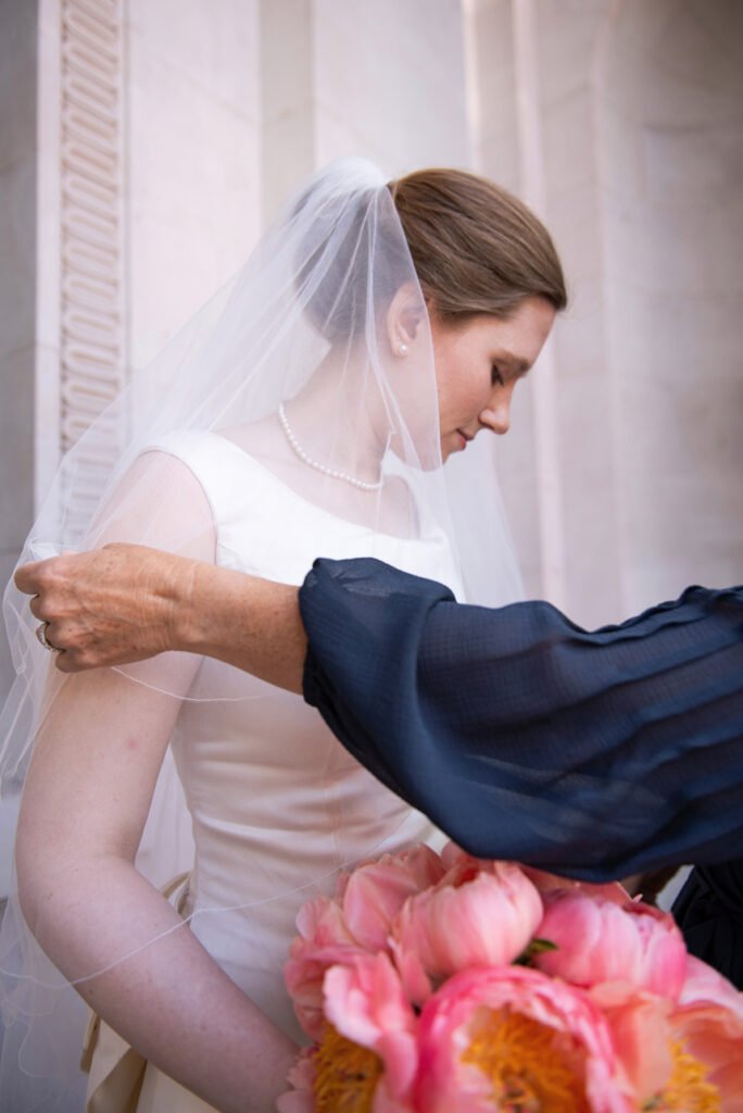 Bridesmaid in navy adjusting the bride's veil beside a bouquet of coral peonies