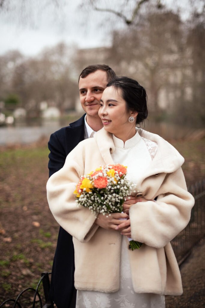 Bride in a cream coat holding an orange bouquet, her groom beside her in a winter park