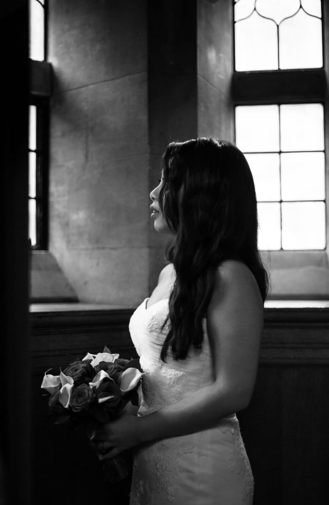 Bride holding a calla lily bouquet beside a tall window, black and white photograph