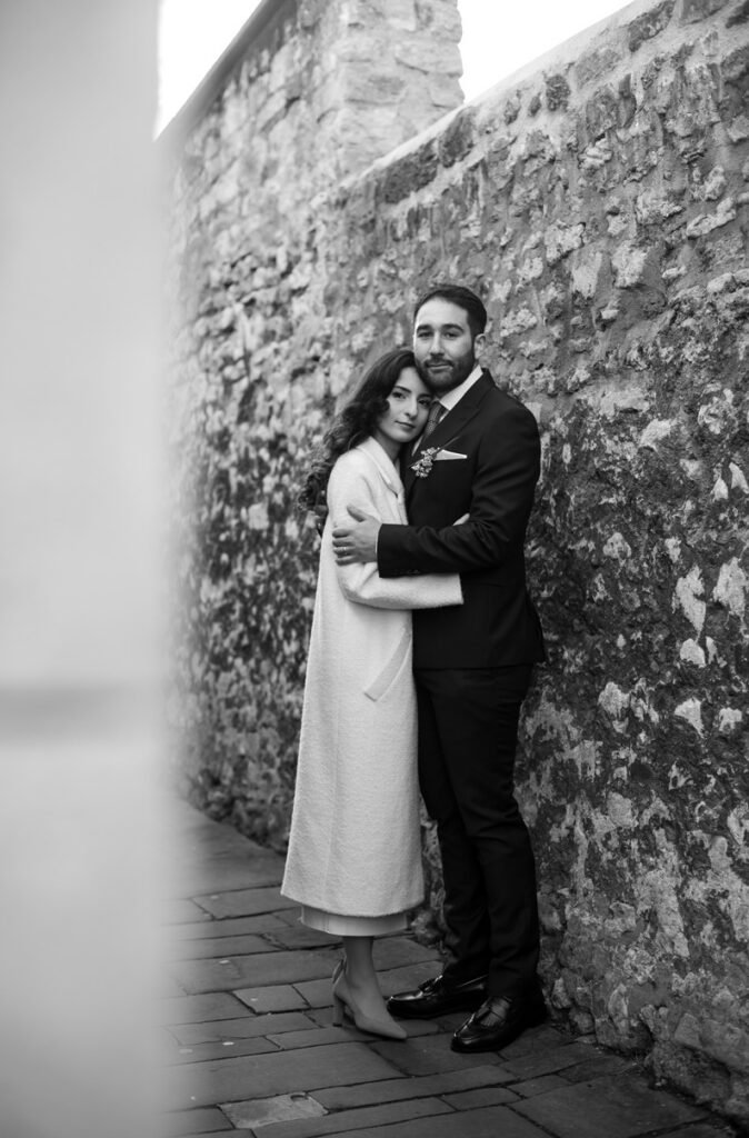 Bride and groom holding each other in a narrow alley beside a flint wall, black and white photograph