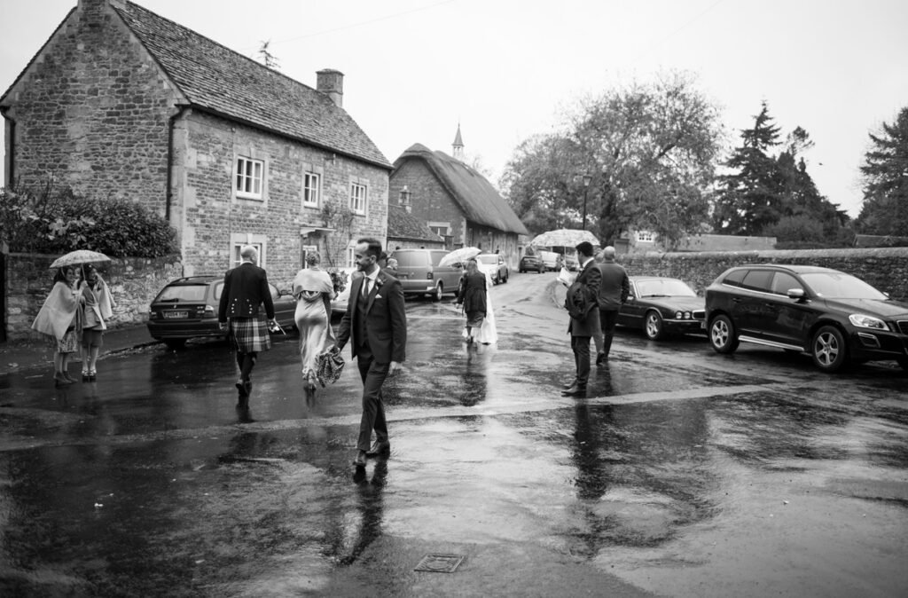 Bride and groom crossing a wet village road under umbrellas, black and white photograph