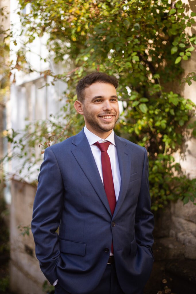 Groom in a navy suit and red tie standing under a climbing rose