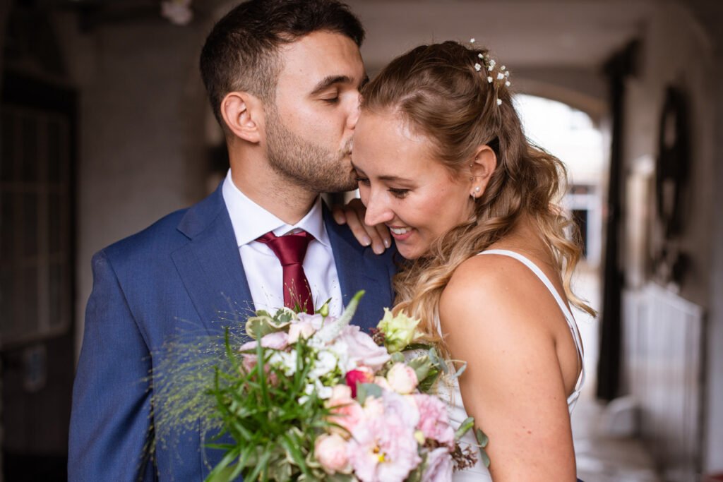 Groom kissing the bride's temple in a stone cloister, bouquet in her arms