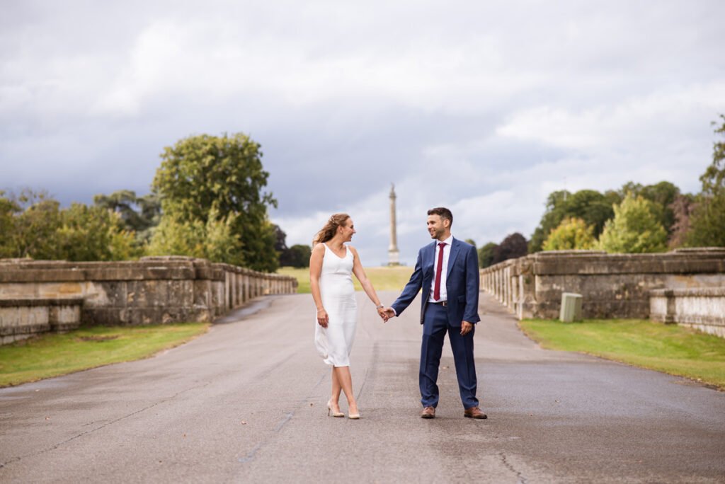 Bride and groom holding hands on a parkland drive, an obelisk in the distance