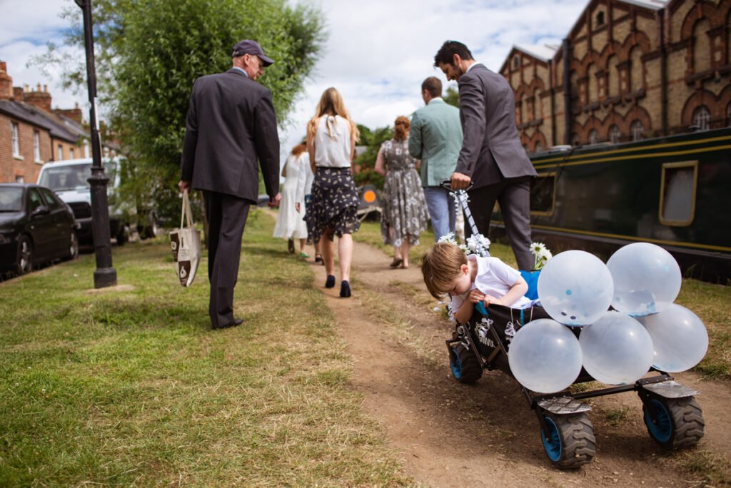 Wedding party walking a riverside path, a child pulling balloons on a cart
