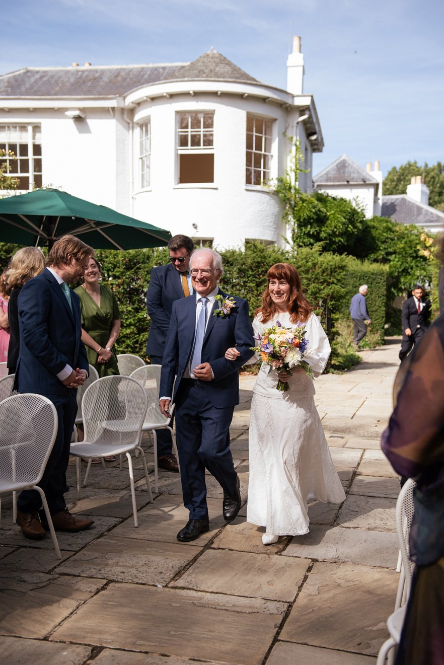 A bride walking down the aisle with her father in Pembroke Lodge garden on the day of her wedding.